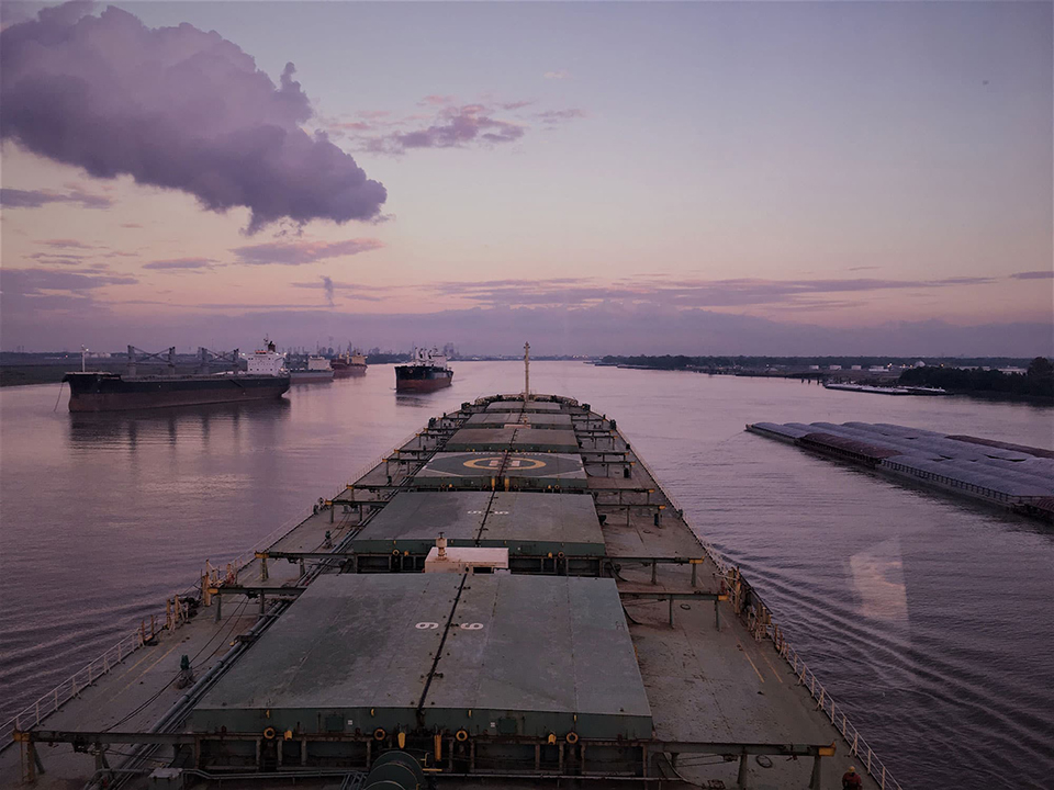View from the M/V <em>Ocean Wind</em> as the ship transits down the Mississippi River with orange, pink, and purple clouds.
