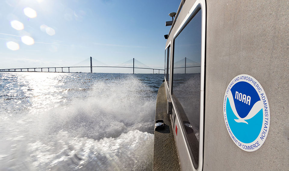 Vessel on the water with a NOAA logo on its side with a view of a bridge in the distance.