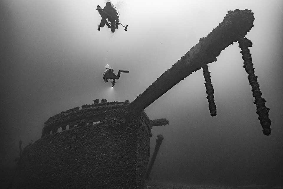 Black and white image of a sunken ship with two people swimming above it.
