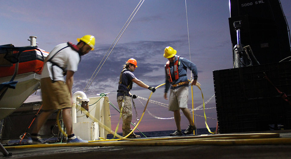 Remotely Operated Vehicle (ROV) technician Jason White (background, left) and Bryan Costa, geospatial scientist, maneuver the ROV over the side of the ship for an evening dive on March 31, 2015. Middlebury College student Ryan McElroy (foreground), an intern on mission, guides the ROV tether along the deck to ensure it doesn't get tangled.