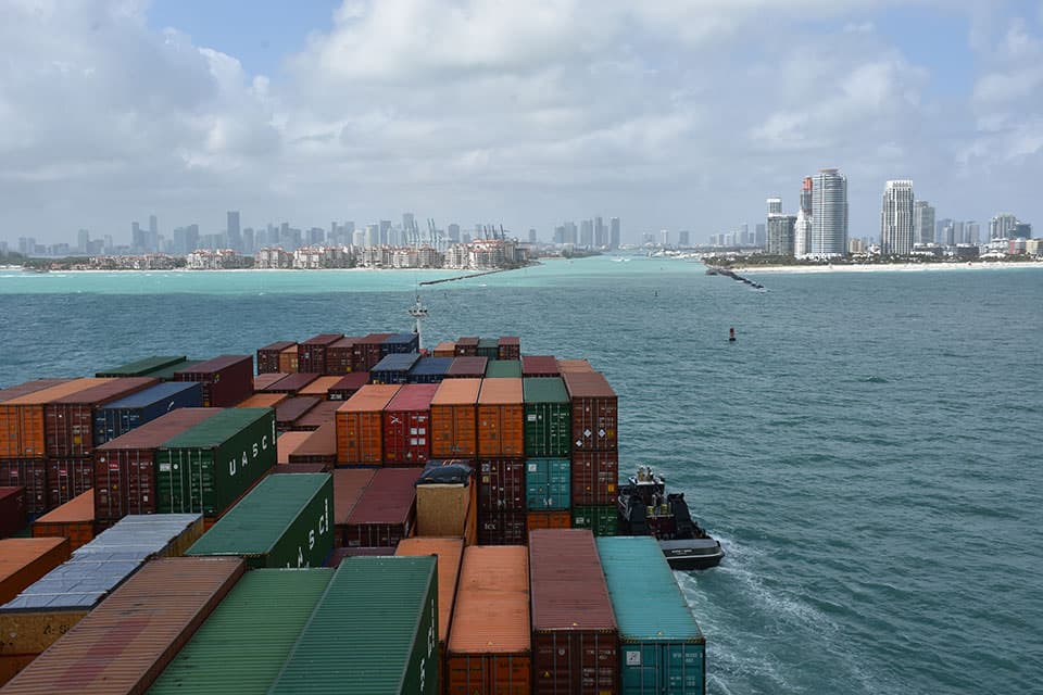 The city of Miami stands in the distance as a shipping vessel carrying multi-colored shipping containers, stacked eleven to fourteen wide and three to four high, is guided into the crystal blue waters of PortMiami by a tug boat.